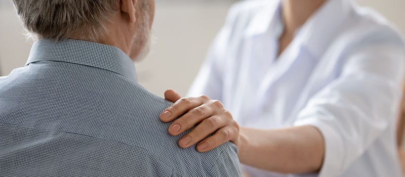 Close up young woman doctor touching senior patient shoulder at meeting in hospital, caring physician gp comforting and supporting mature man at medical appointment