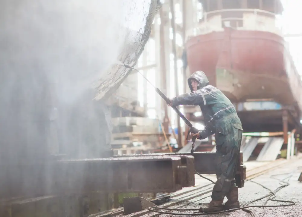 Worker using a high-pressure hose on a boat inside a shipyard workshop.